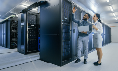 Two IT professionals in a data center discussing server infrastructure, with one holding a laptop and pointing at a server rack