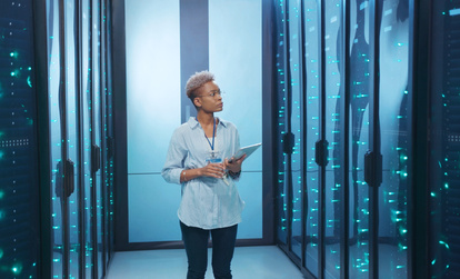 A female IT professional holding a tablet stands in a server room surrounded by illuminated data servers with glowing blue lights