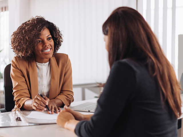 Two professional women engaged in a business meeting in a modern office space, one smiling and holding a pen while discussing documents on the table