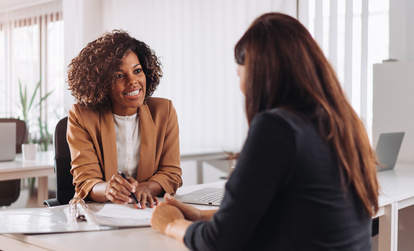 Two professional women engaged in a business meeting in a modern office space, one smiling and holding a pen while discussing documents on the table