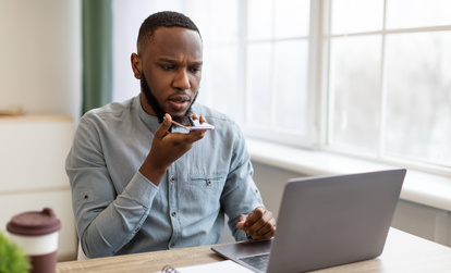 A young man sitting at a desk with a laptop, speaking into his smartphone with a focused expression, while a notebook and coffee cup are nearby